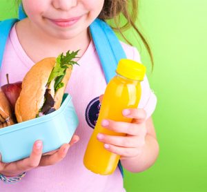 Young schoolgirl with lunch box on a green background. Little girl with a school backpack and a set of food for a snack. Free space for text