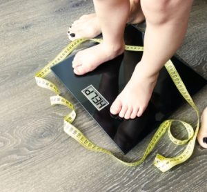 The feet of a child standing on a weighing scale with the LCD depicting the word "help". A parent stands behind.
